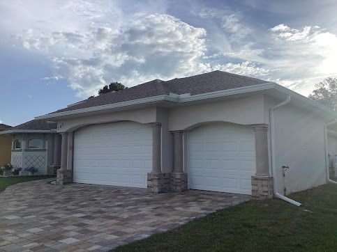 Two-car garage with white doors, beige facade, and brick paved driveway under cloudy sky.