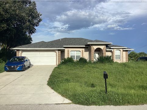 House with overgrown grass; blue car in driveway. Cloudy sky overhead.