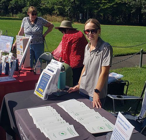Three women at an outdoor information table with brochures. One woman stands behind the table.