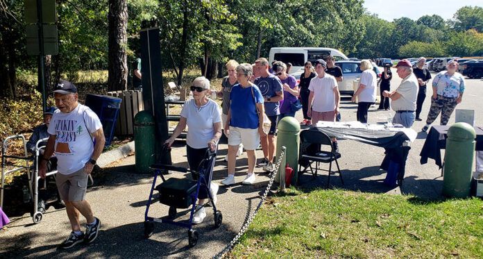 People gather near a table outdoors with some using walkers and other mobility aids. Sunny day.