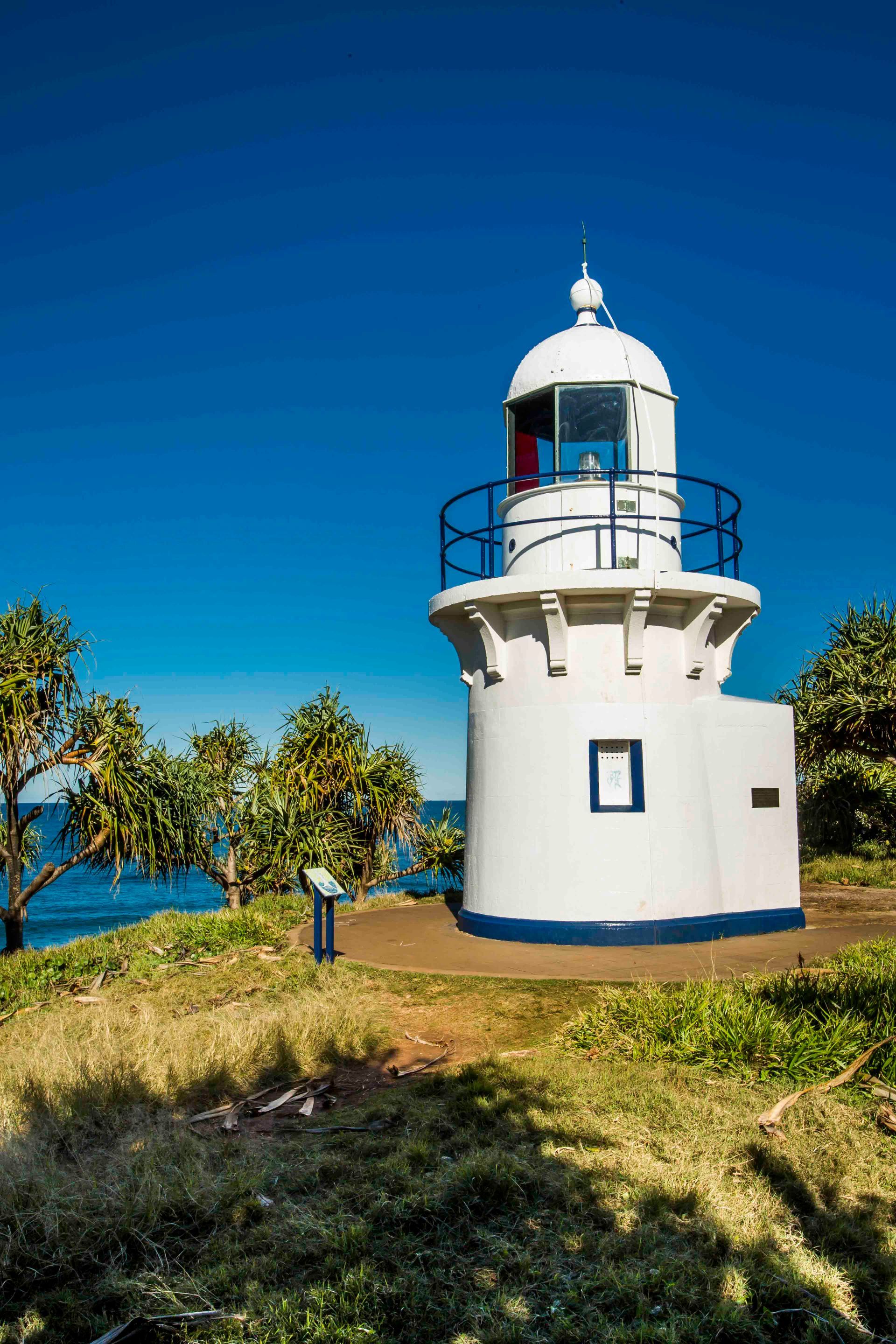 A White Lighthouse Is Sitting On Top Of A Grassy Hill Next To The Ocean — Tweed Byron Recycling In Tweed Heads, NSW