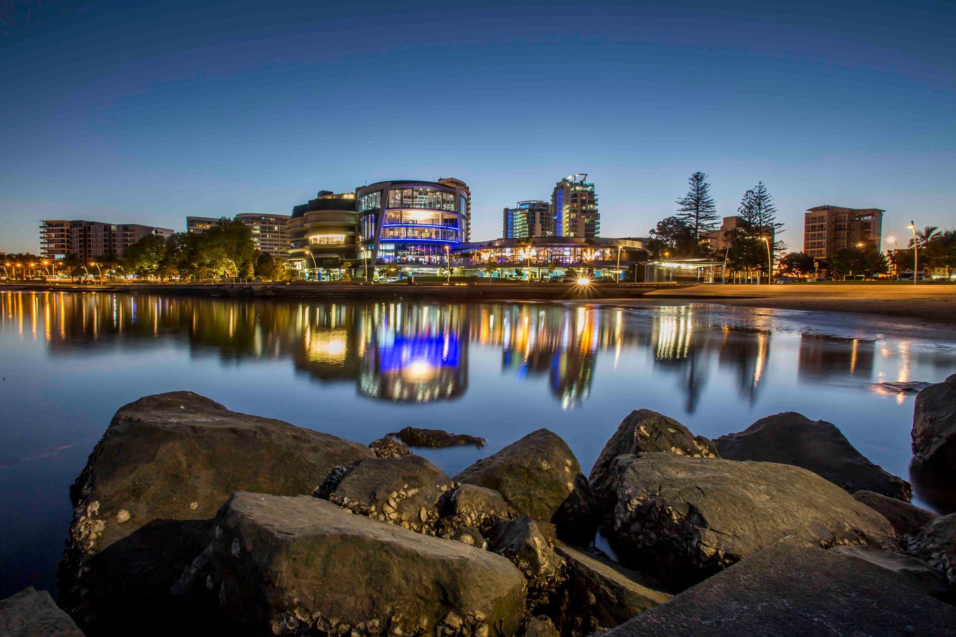 A City Skyline Is Reflected In A Body Of Water With Rocks In The Foreground — Tweed Byron Recycling In Tweed Heads, NSW