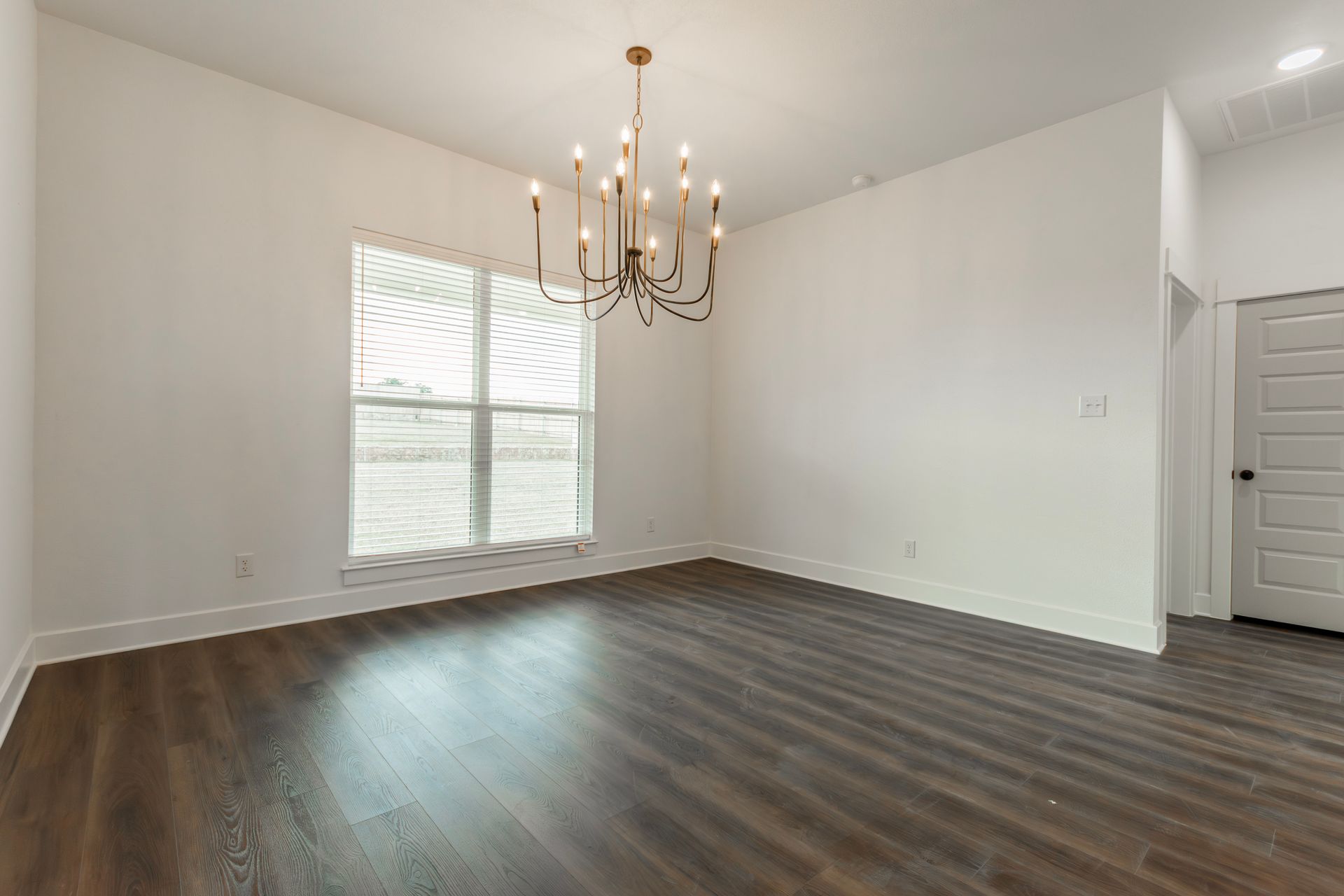 Empty dining room with dark wood flooring, white walls, large window, and chandelier.