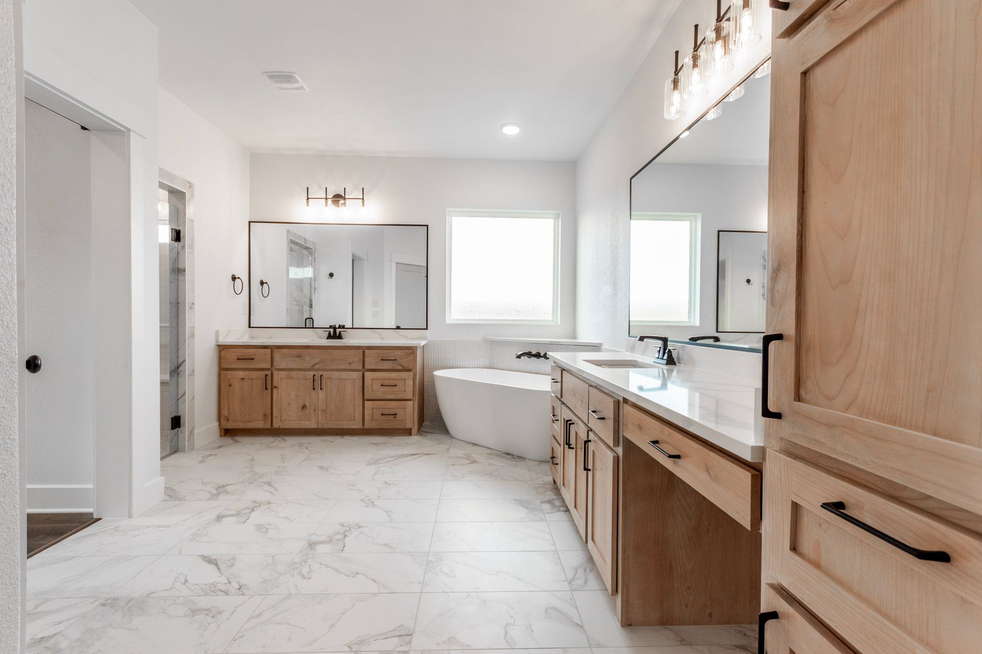 Spacious, light-filled bathroom with wood cabinets, large mirrors, and white marble-look flooring.
