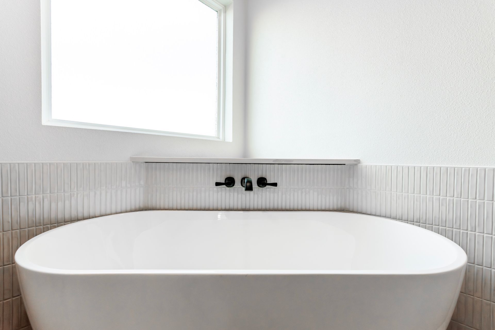 White bathtub with black fixtures, set against white tile wall, near a window.