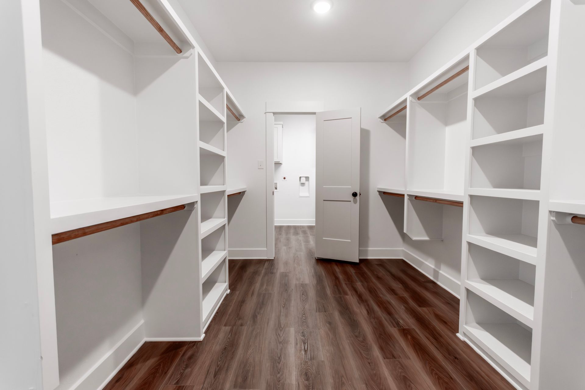 Empty white walk-in closet with shelves and hanging rods, wooden floor, and a door to another room.