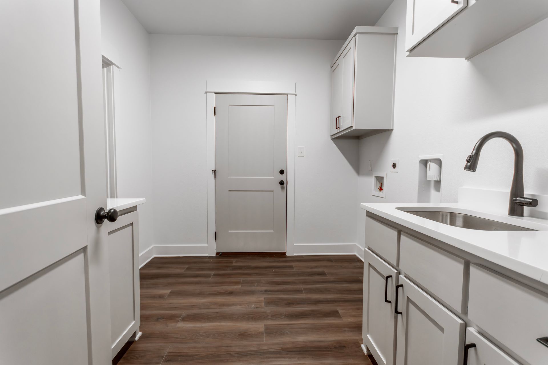 Laundry room with white cabinets, sink, and wood-look flooring. White door on wall.