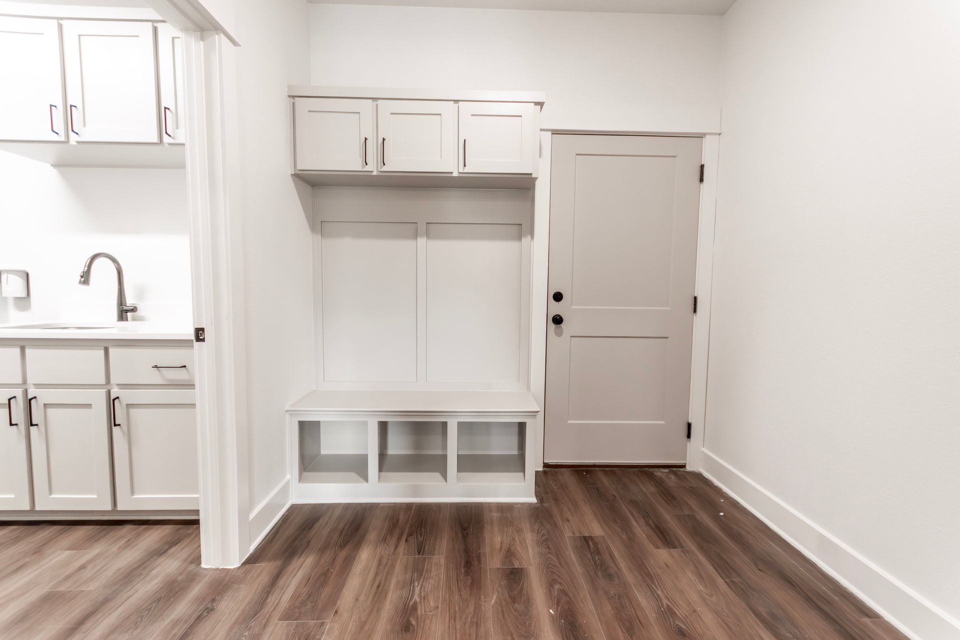 Mudroom with built-in bench, cubbies, and overhead cabinets; white walls and door, and wood-look flooring.