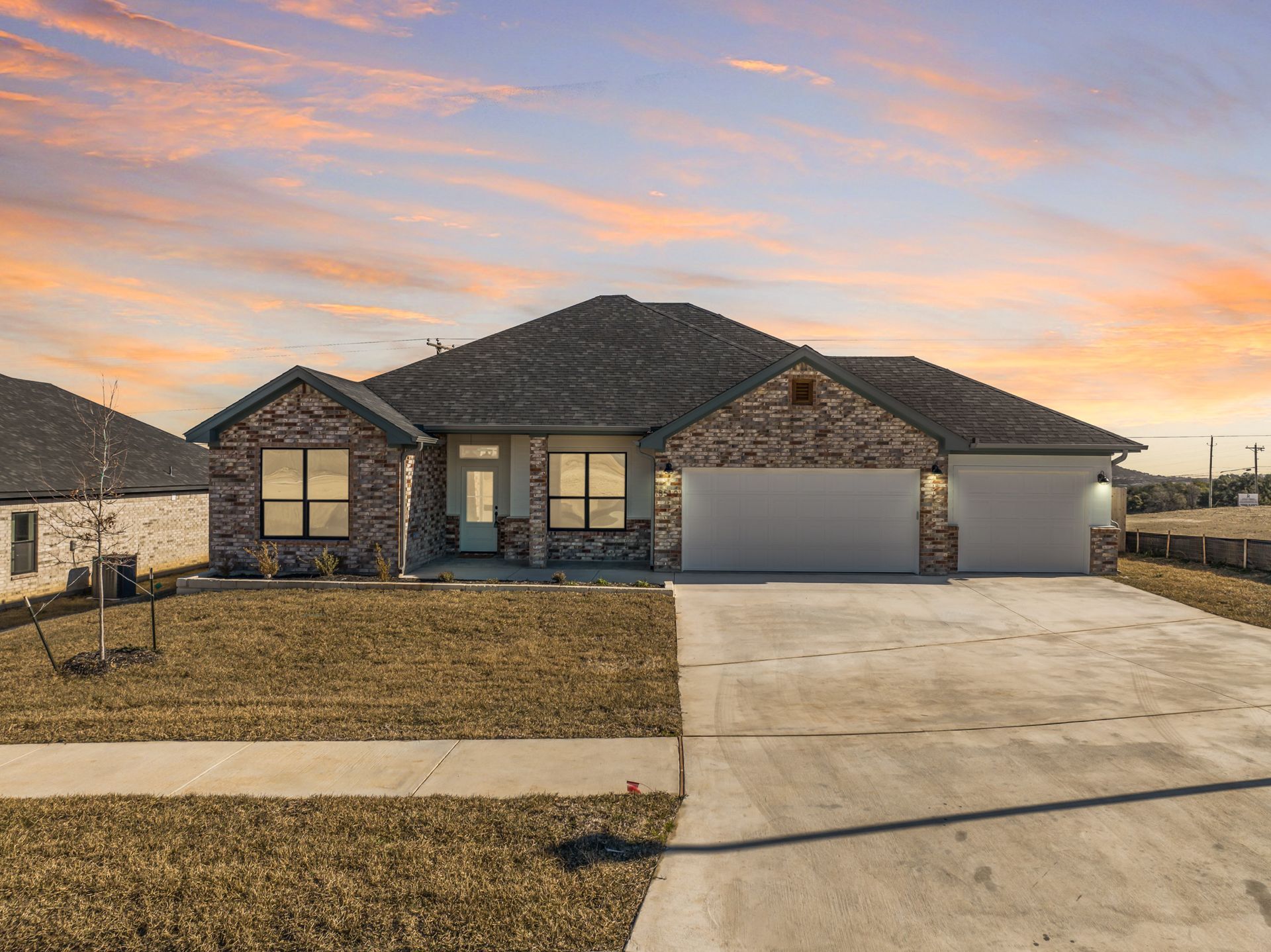 A single-story, brick-facade suburban house with a three-car garage and a concrete driveway at sunset.