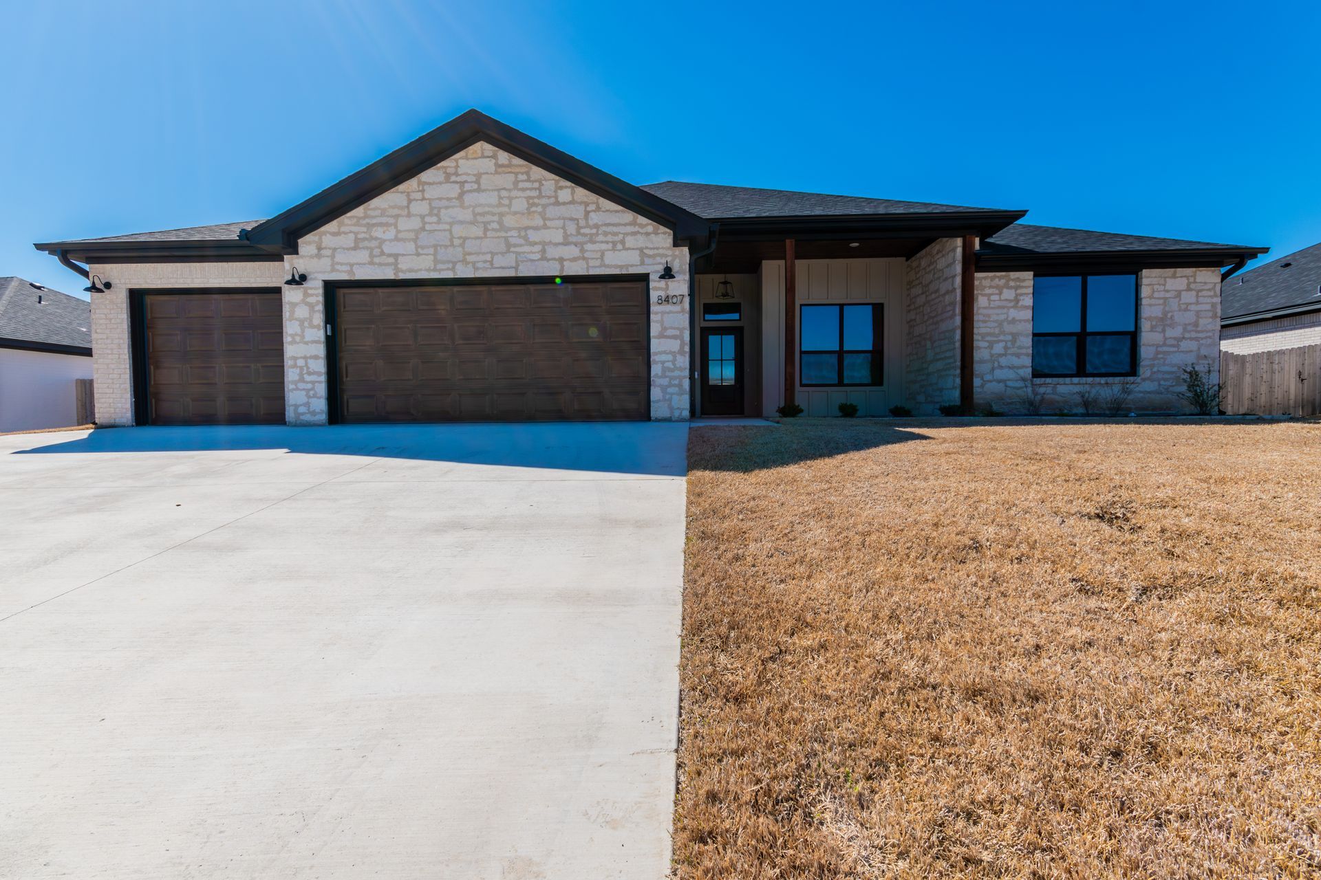 A single-story suburban house with stone siding, a dark roof, a two-car garage, and a concrete driveway under a blue sky.