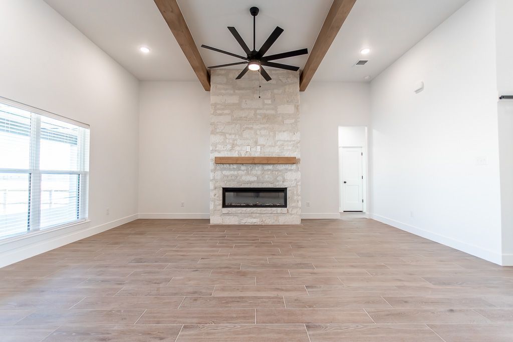 Empty living room with fireplace, wooden beams, light wood floors, and ceiling fan.