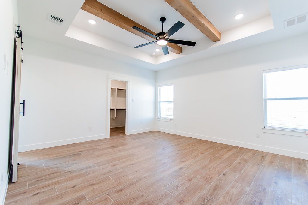 Empty bedroom with wood-look flooring, white walls, walk-in closet, and wooden ceiling beams.