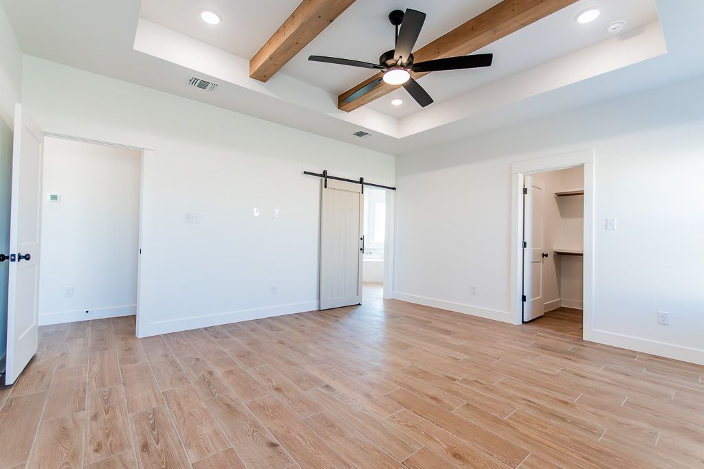 Empty, modern bedroom with wood-look tile floor, white walls, exposed ceiling beams, ceiling fan, and sliding barn door.
