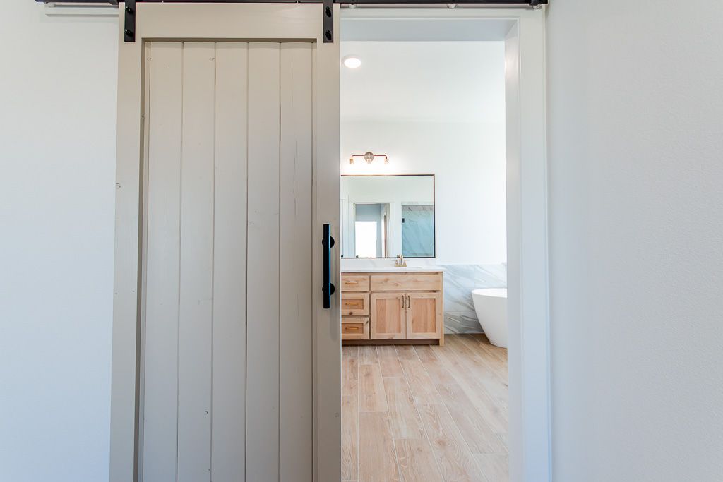 Gray barn door partially open revealing a bathroom with wood vanity, large mirror, and soaking tub.