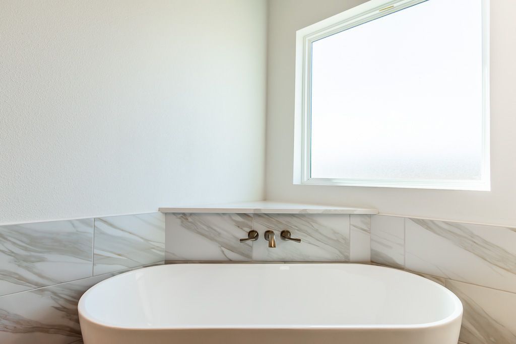 Freestanding white bathtub below a window in a corner, with marble tile accents.