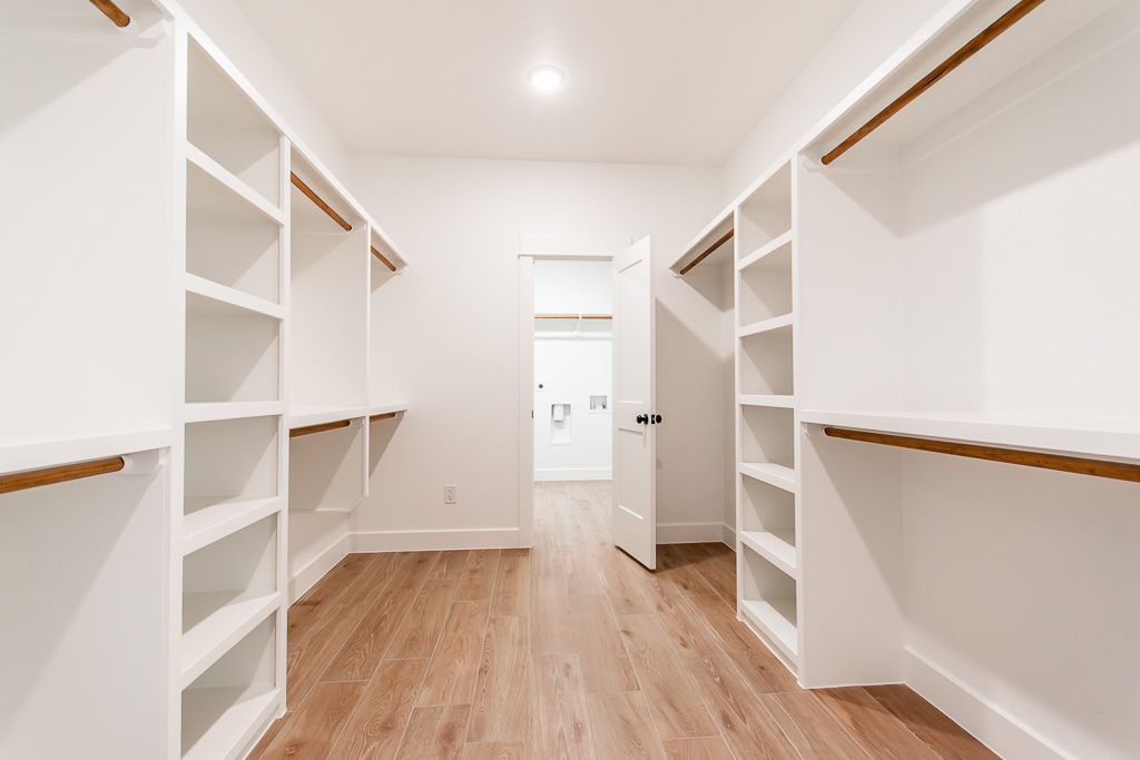 Empty white walk-in closet with shelves and hanging rods, wooden floor, and a door to another room.