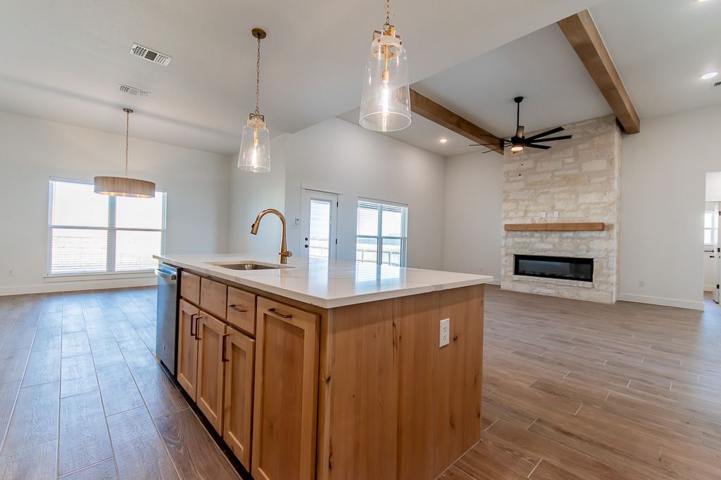 Open-concept kitchen with island, pendant lights, and fireplace with wooden beams and light wood floors.
