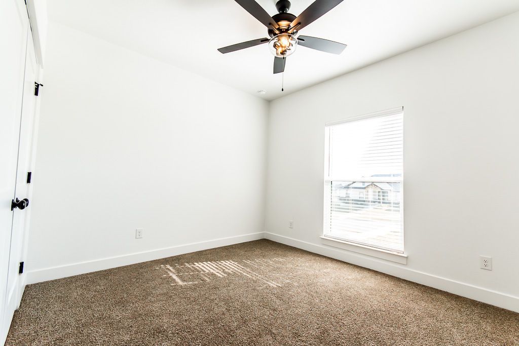 Empty bedroom with white walls, brown carpet, window, and ceiling fan.