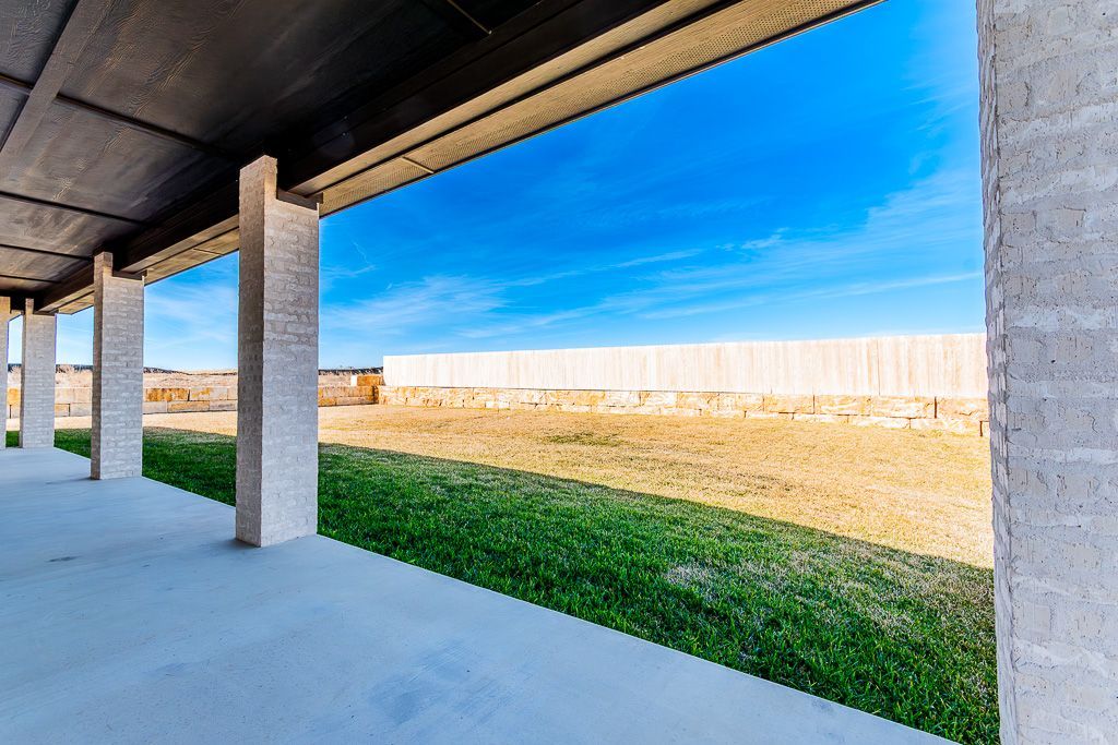 Concrete patio with columns looking out onto a grassy field and a wooden fence under a blue sky.