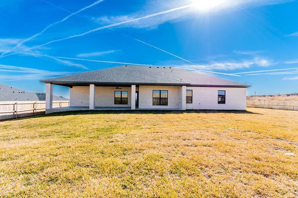 Rear view of a light-colored single-story house with a covered patio, set in a grassy field under a bright blue sky.
