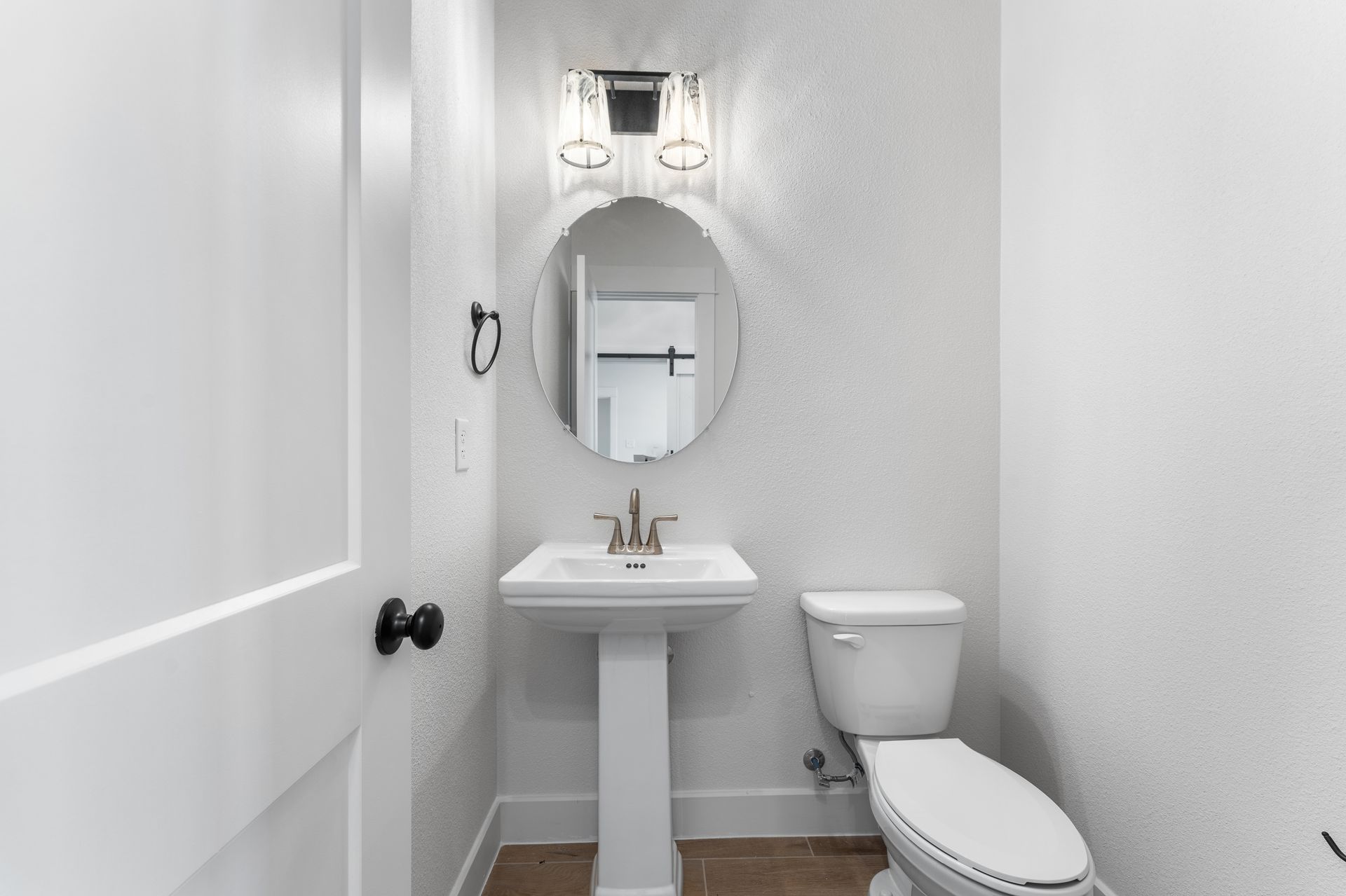 Small white bathroom with pedestal sink, oval mirror, and toilet. Black door knob and towel ring.