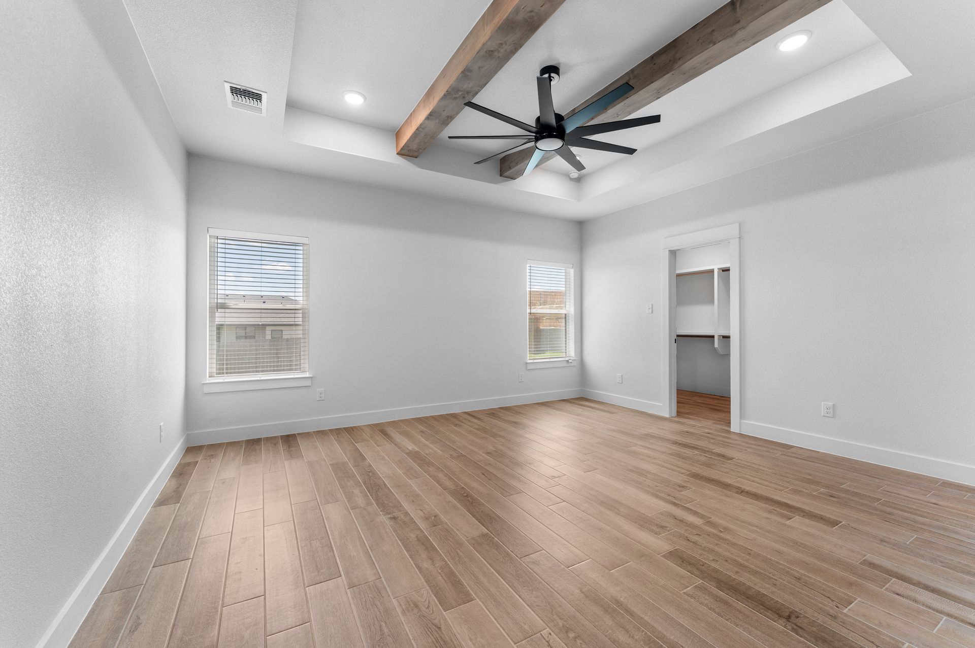 Empty bedroom with wood floors, white walls, recessed ceiling, and two windows.