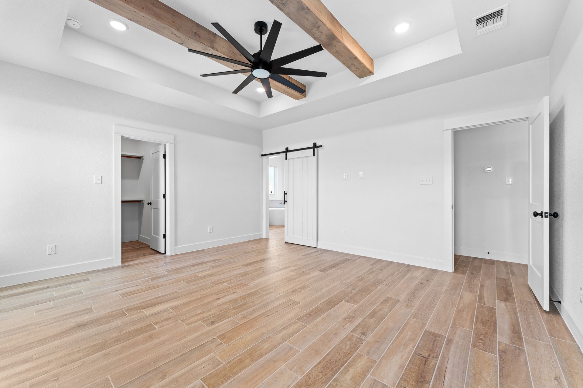 Spacious white bedroom with wood floors and beams, a ceiling fan, and open doorways.