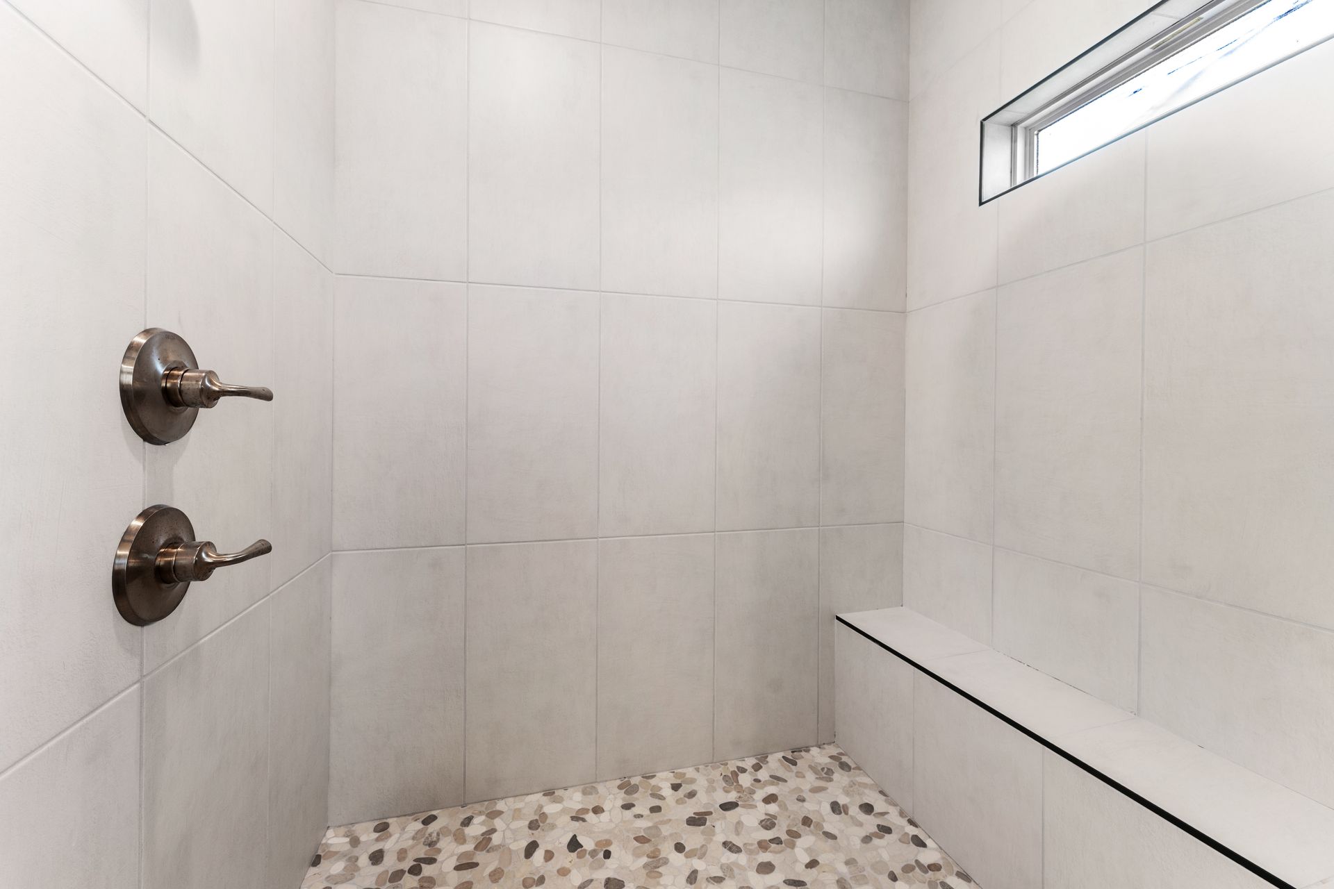Shower with white tiled walls, pebble floor, bench, and metal fixtures.