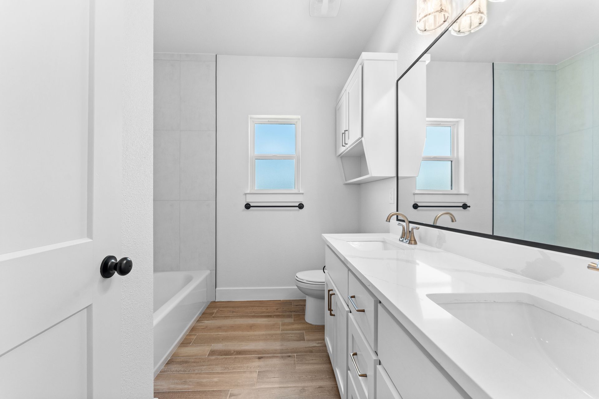 White bathroom with long vanity, toilet, tub, small window, and hardwood floor.