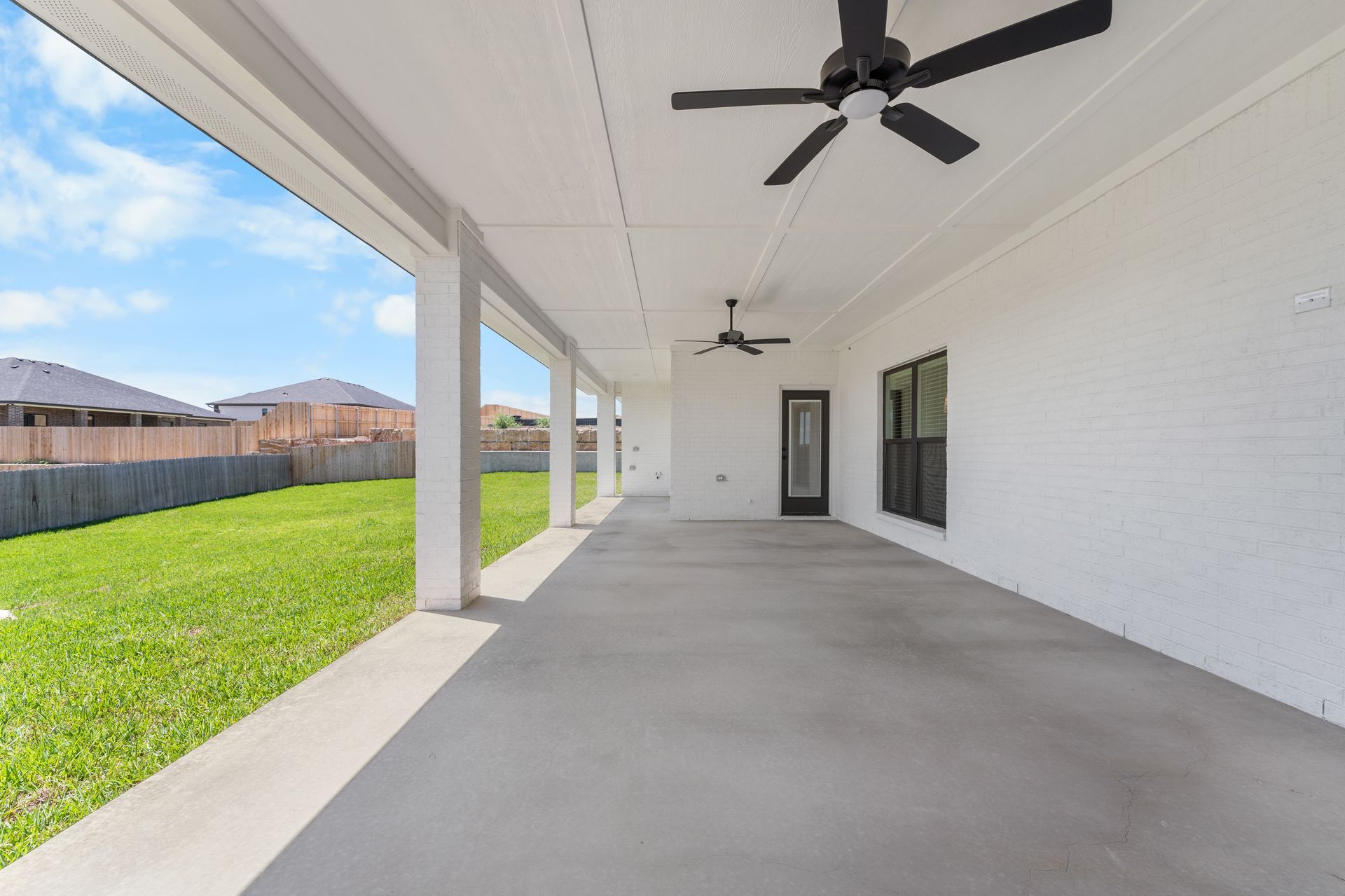 Spacious covered patio with ceiling fans, concrete floor, brick walls, and a grassy backyard under a blue sky.