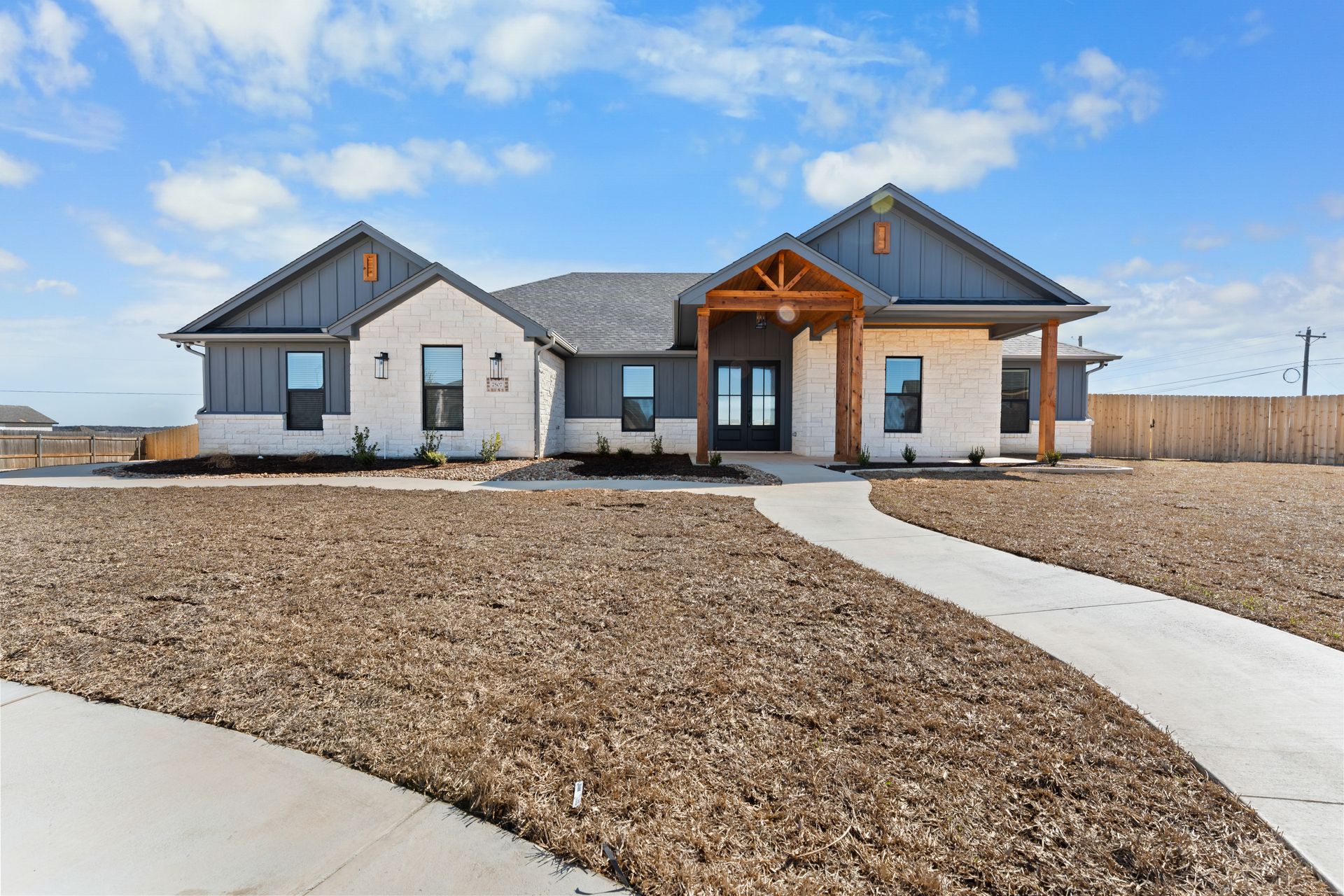A modern, one-story house with white stone and grey siding under a blue sky, featuring a wooden porch and paved walkway.