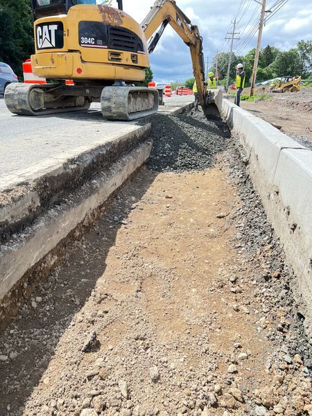 A yellow excavator works on a road, removing materials from a trench beside a new concrete curb.
