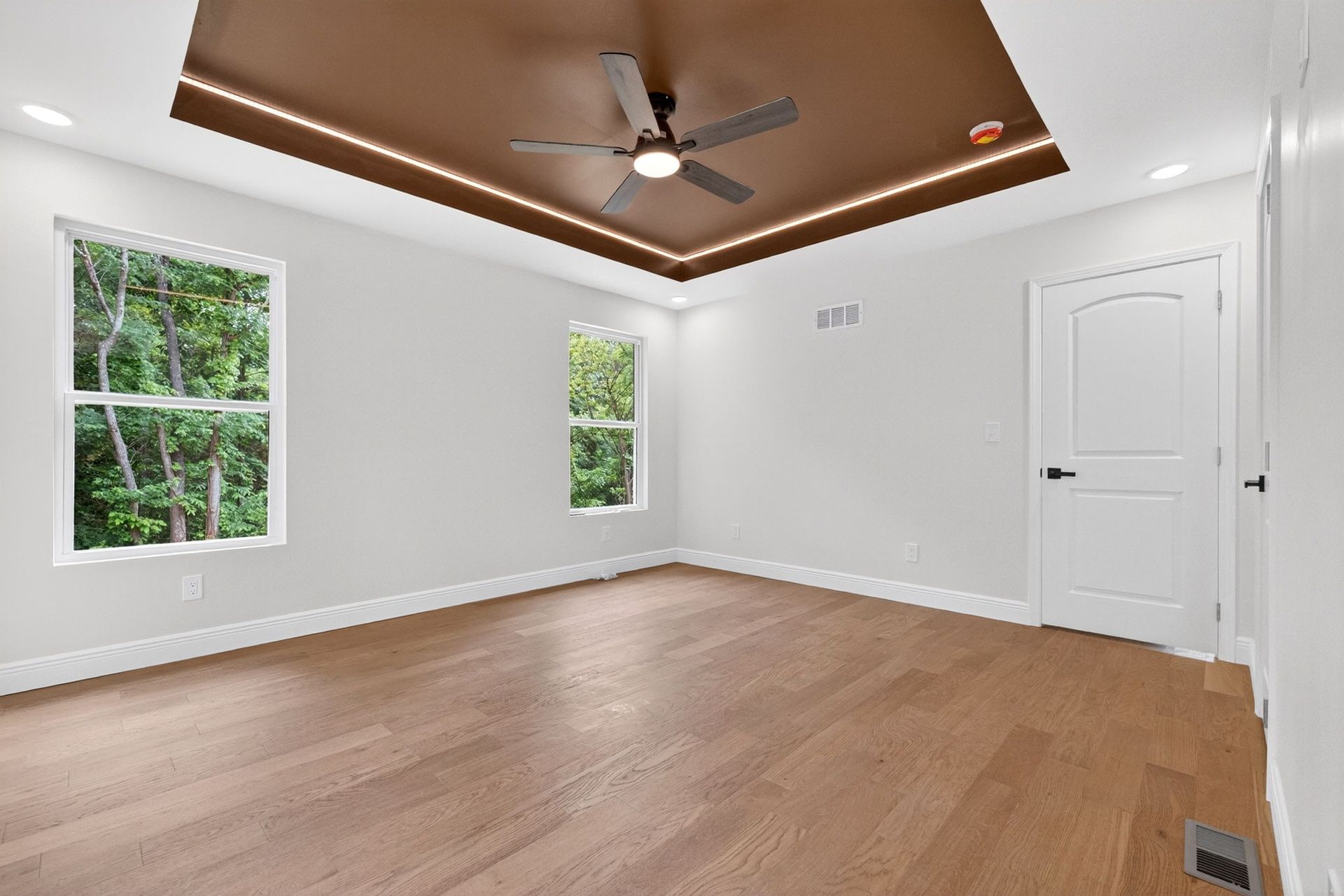 Empty white bedroom with hardwood floors, tray ceiling with ceiling fan, and three windows