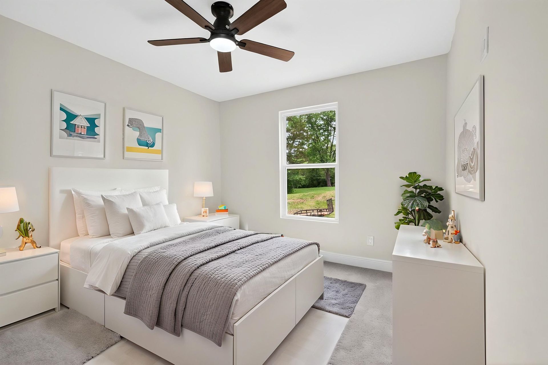 Bright bedroom with a bed, white walls, ceiling fan, and a window with plants and framed art