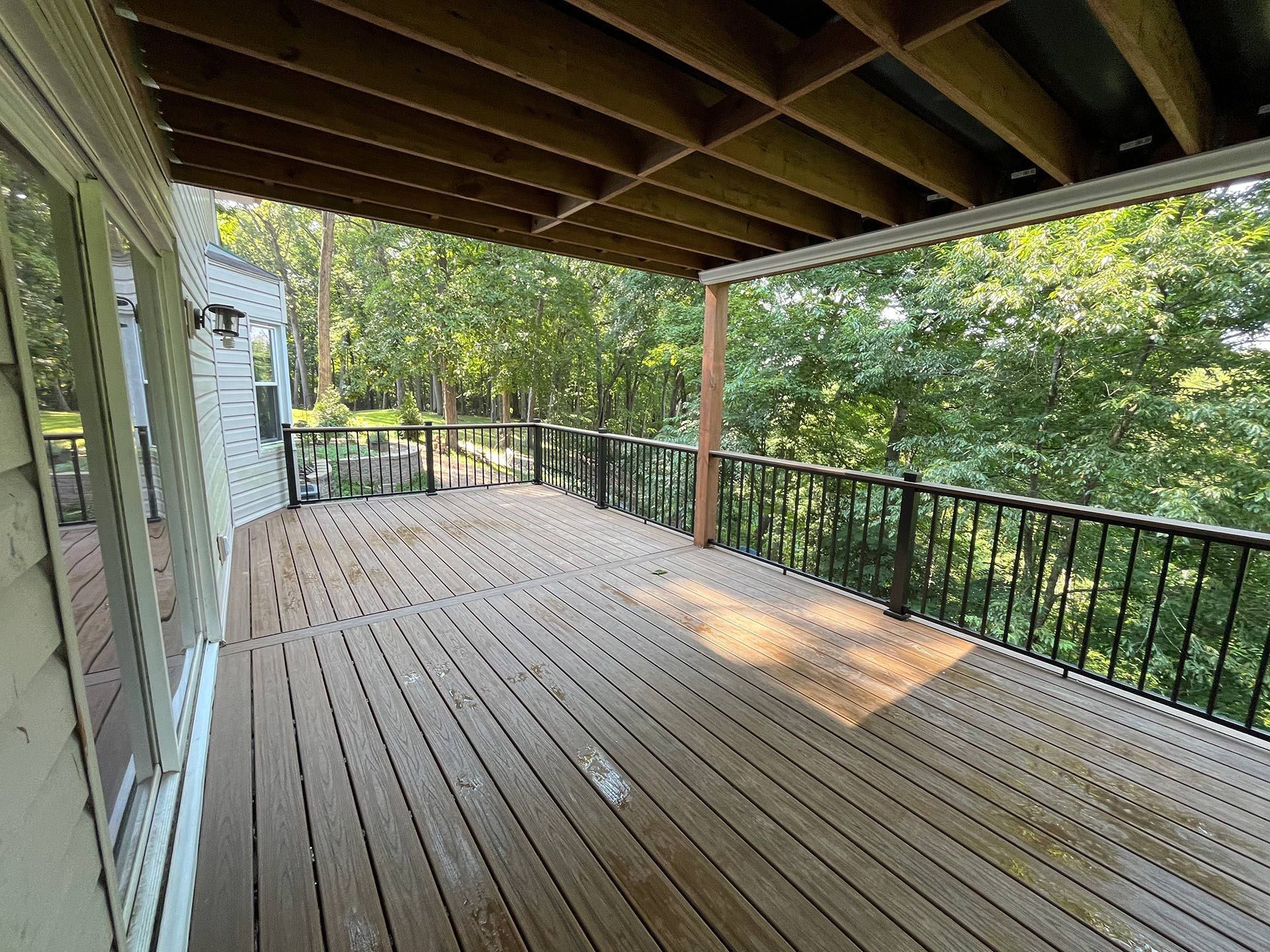Covered wooden deck with black railing, trees beyond, and a house wall on the left.