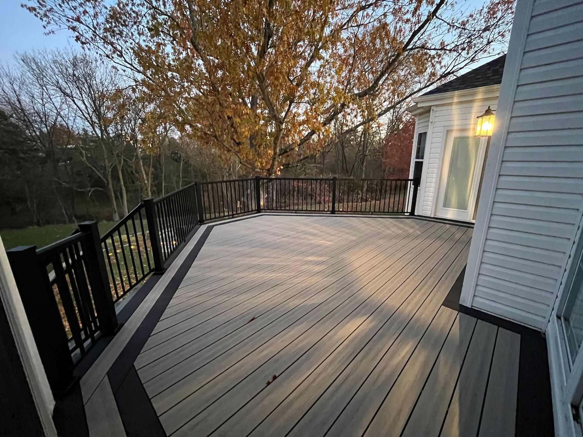 Spacious wooden deck with black railing beside a house, overlooking trees at sunset.