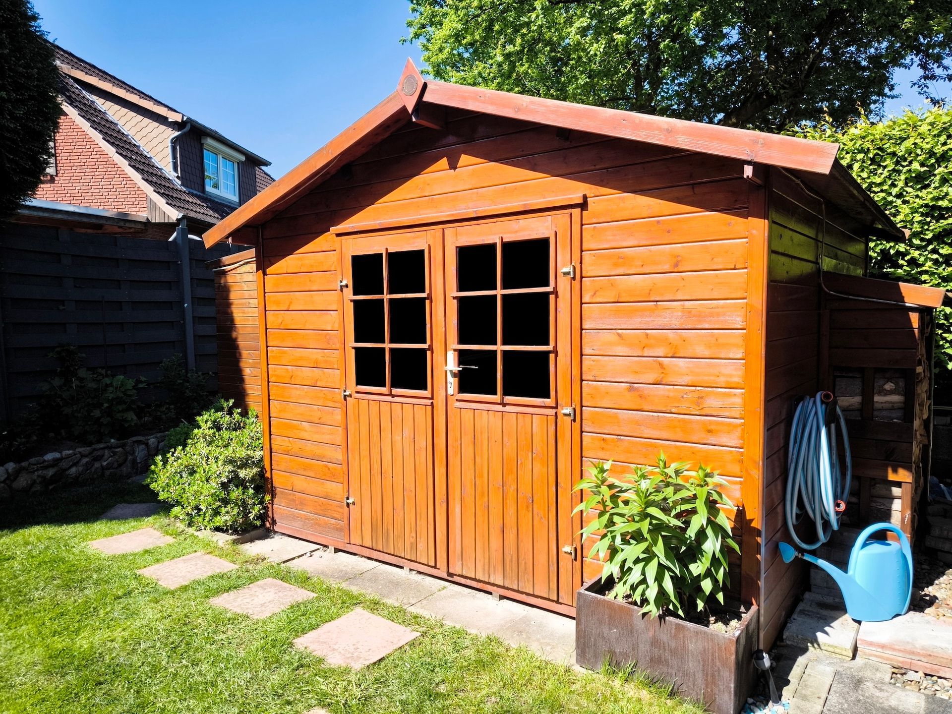 Wooden garden shed with double doors, windows, and a blue watering can beside it in a sunny yard