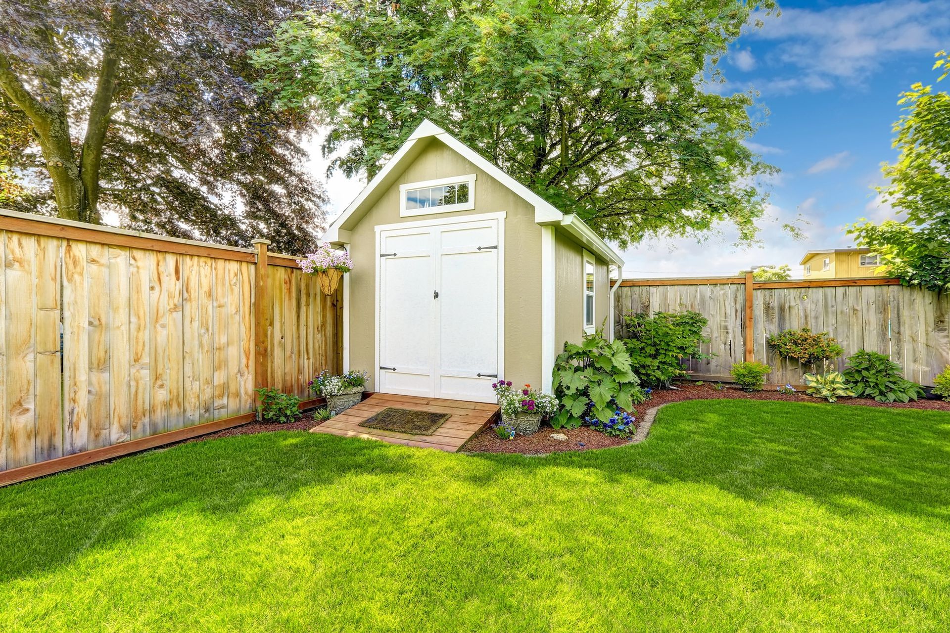 Small white garden shed with double doors in a green backyard, bordered by wooden fences and trees.