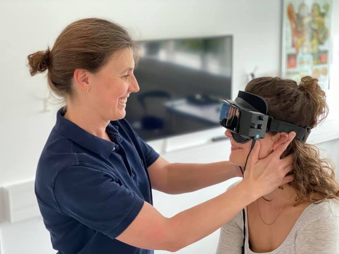Kvinde monterer VR-headset på en anden person indendørs. Begge smiler hos Charlottenlund privathospital.