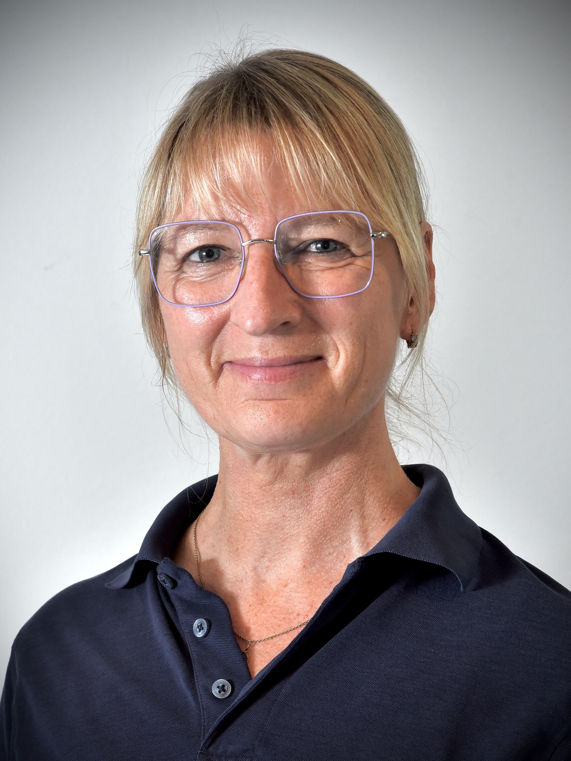 Kvinna med blont hår och glasögon, leende, klädd i blå skjorta, mot en vit bakgrund.