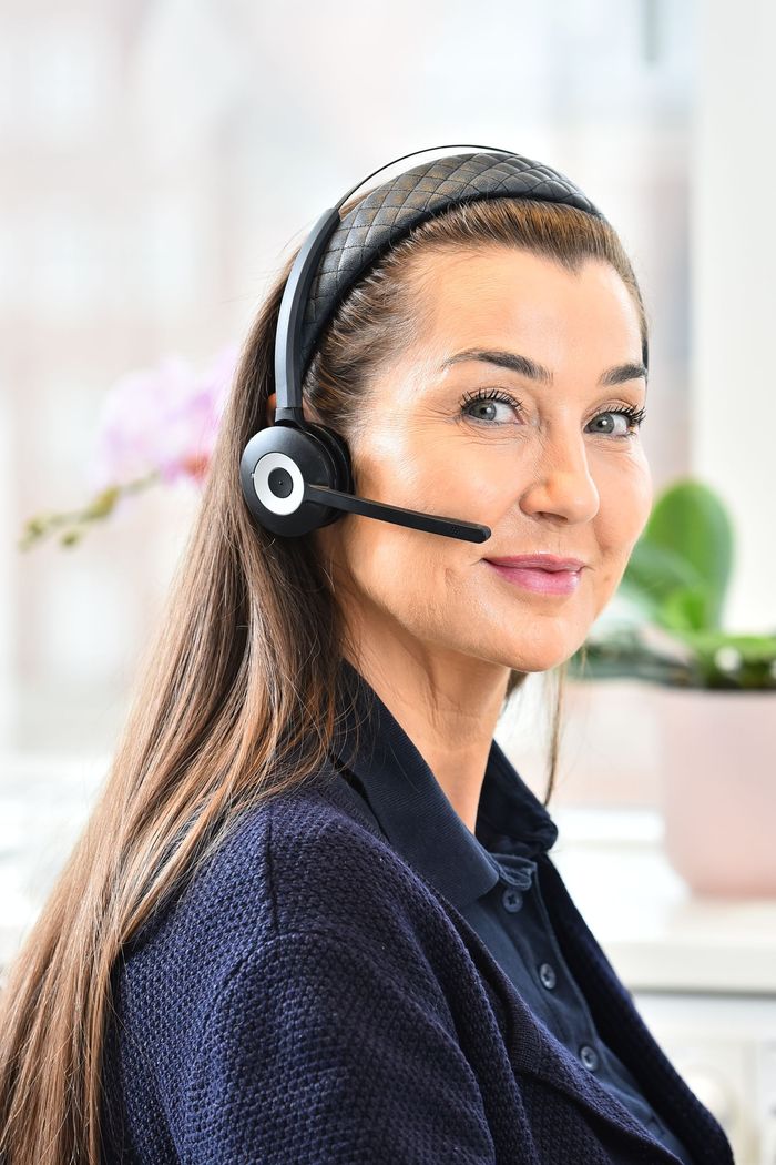 Kvinde med headset, smiler til kameraet, indendørs nær et vindue med en plante i baggrunden, hos Charlottenlund privathospital.