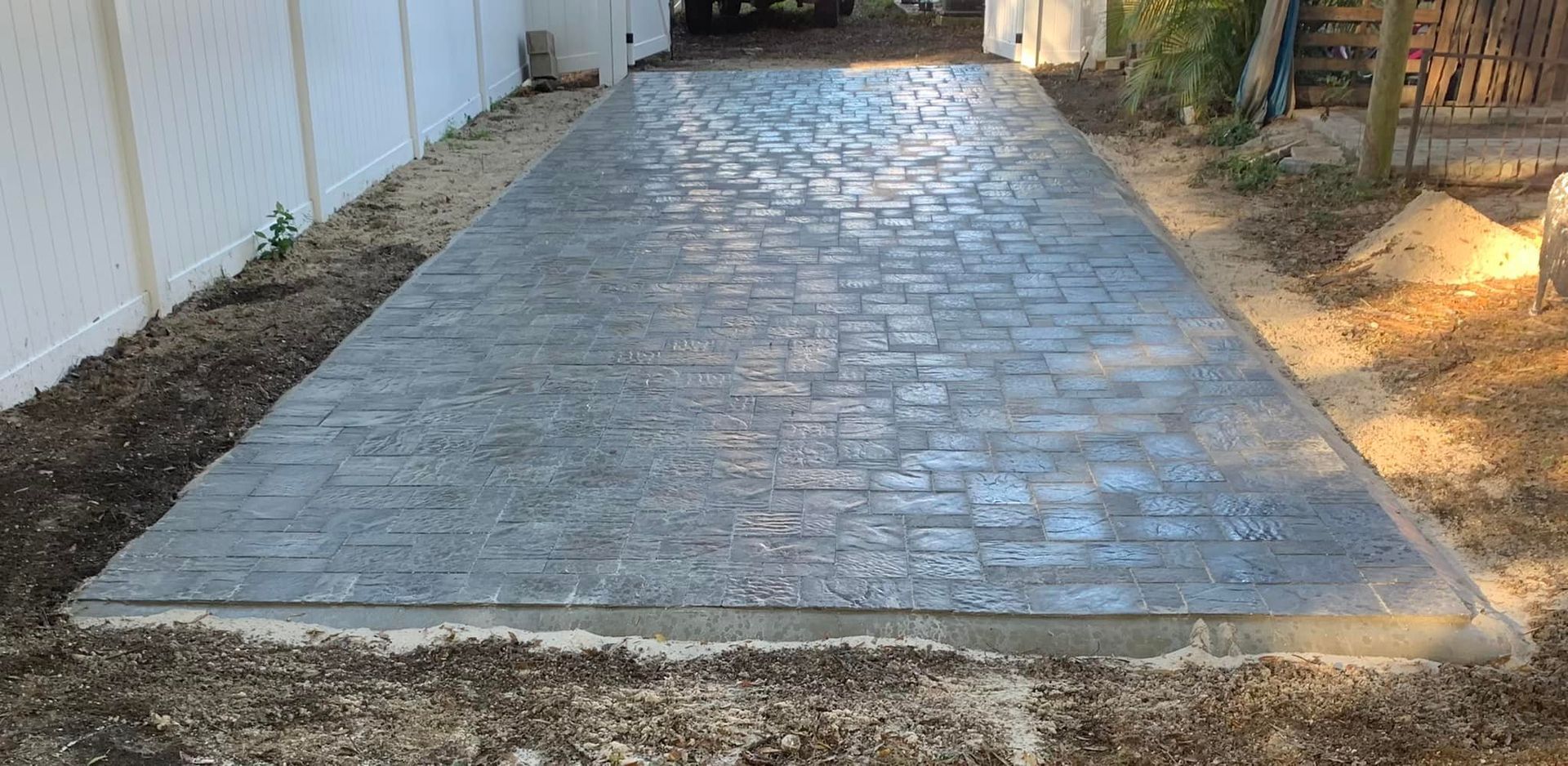 Driveway with brick-patterned concrete between a white fence and dirt.