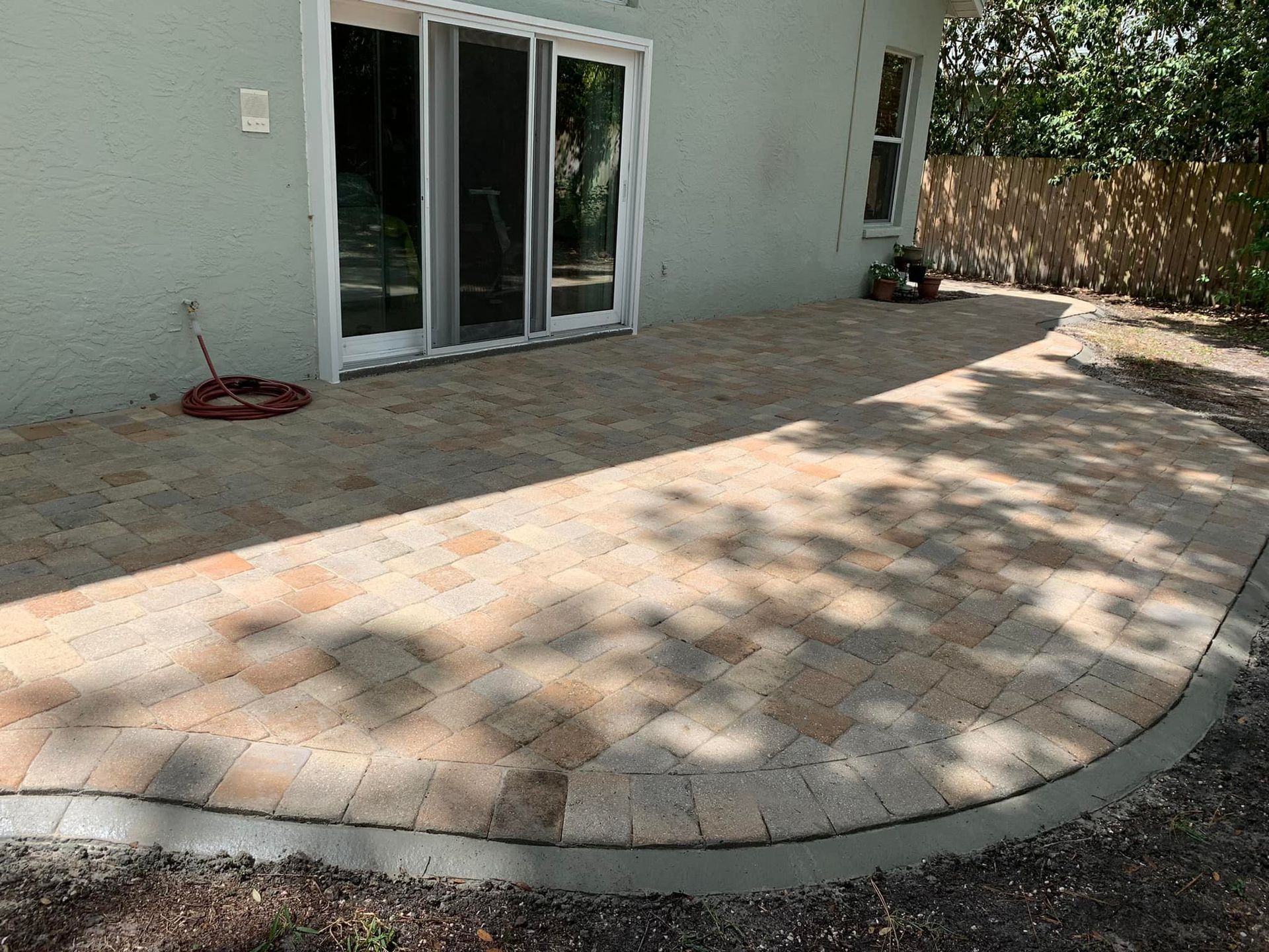A brick patio outside a light green house with sliding glass doors, edged by a concrete border.