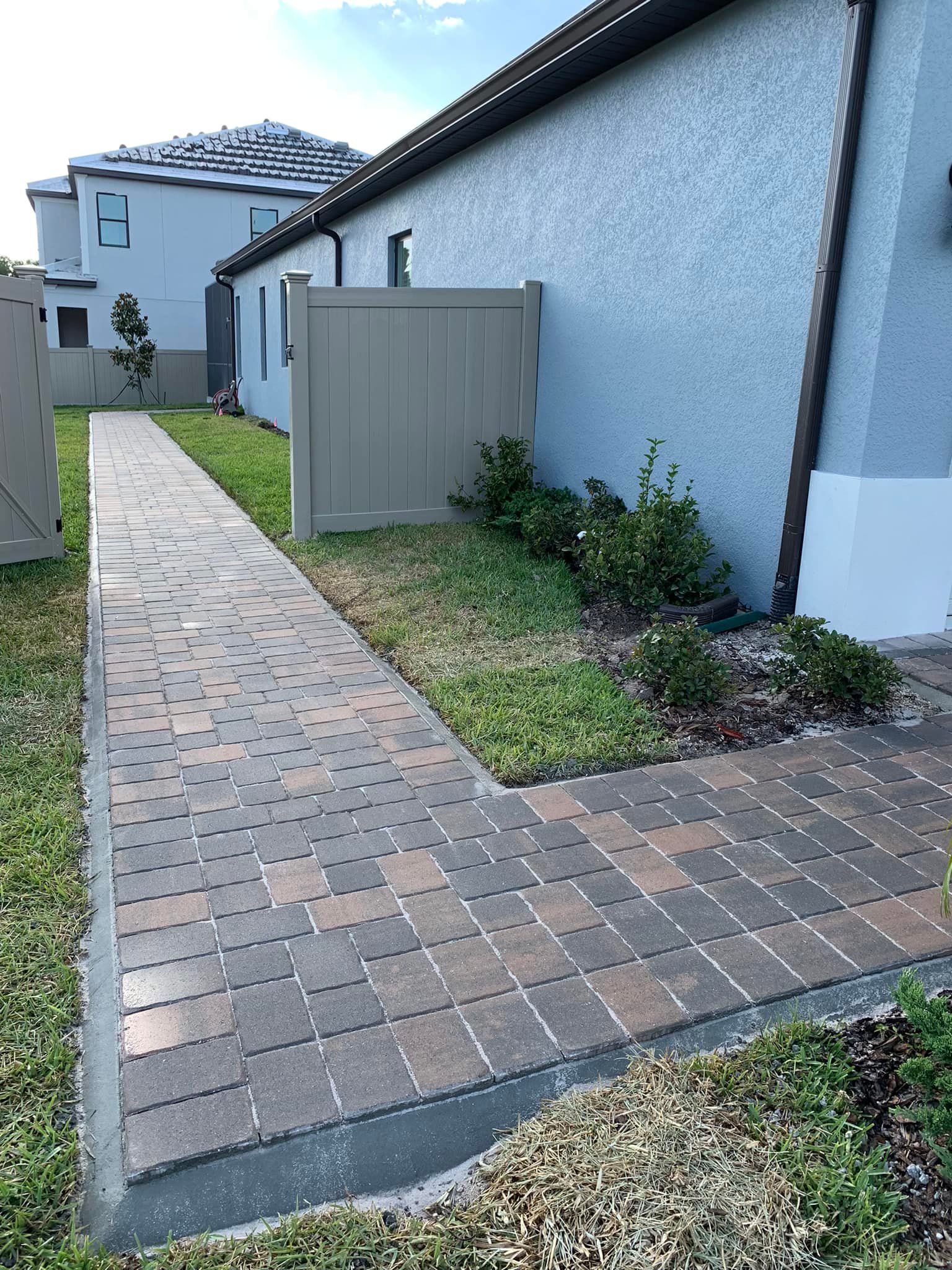 Brick walkway beside a stucco building, with grass, bushes, and a fence.