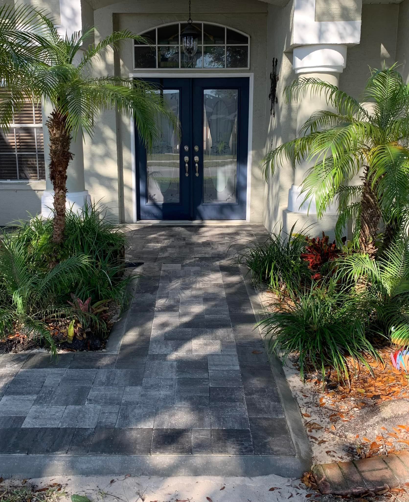 Brick path leading to blue double doors of a house, flanked by greenery.