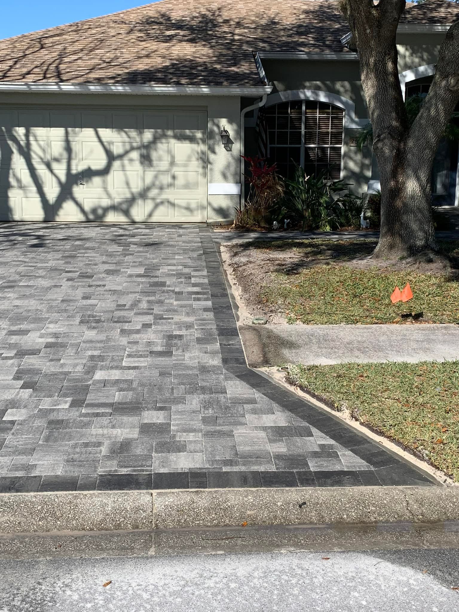 Paver driveway in front of a house, bordered by grass and a tree; gray and black tones.