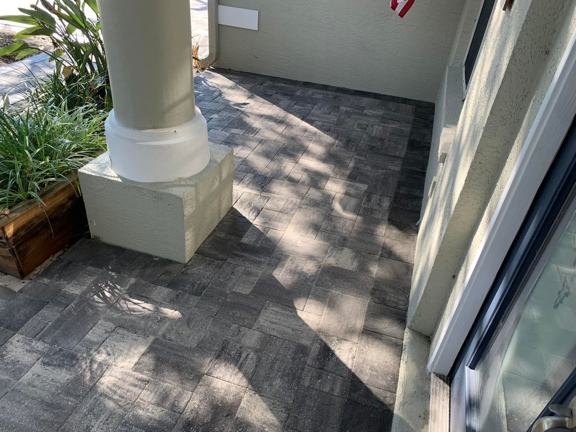 Gray brick pavers on a porch near a pillar and a window. Sunlight creates long shadows.