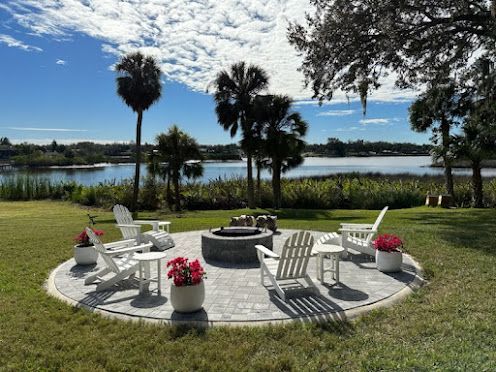 Fire pit with a burning fire at night. Two white chairs nearby. Water and lights in the background.