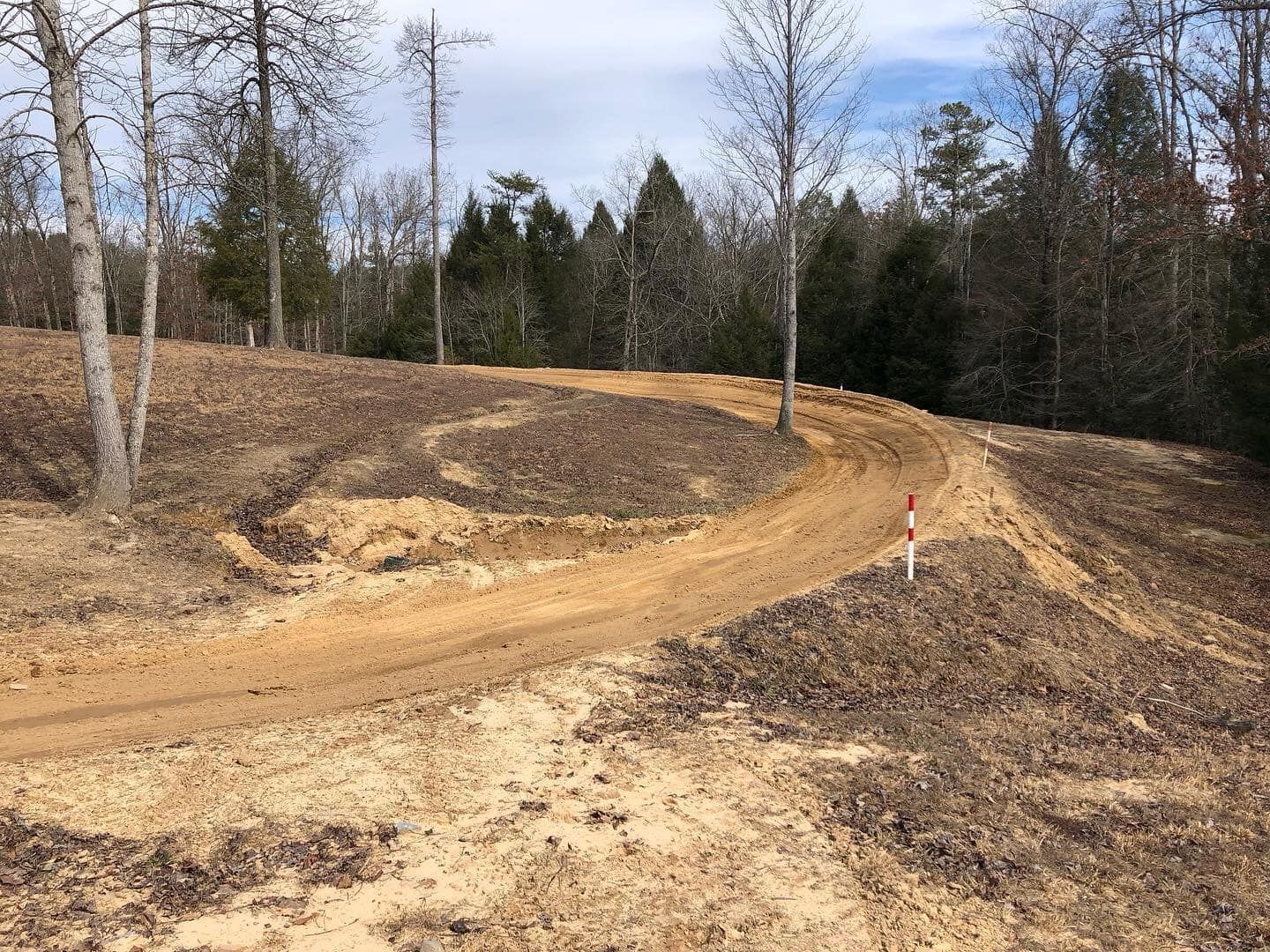 A dirt road winds through a cleared, wooded area with brown fallen leaves and a solitary red marker stake.