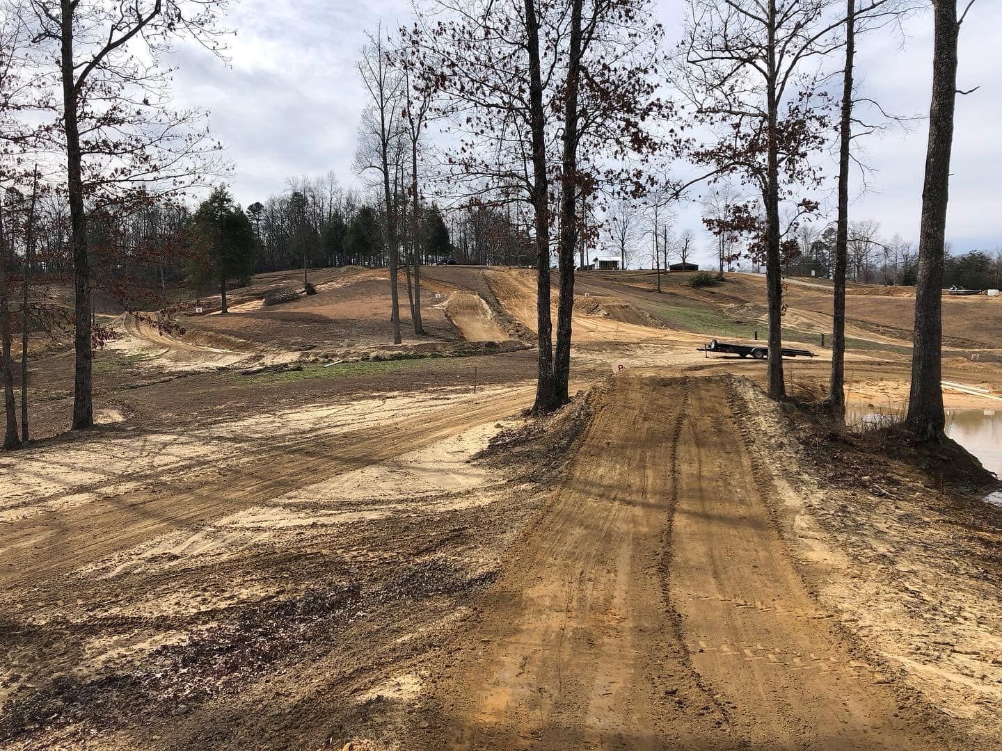 A dirt path leads through a cleared, wooded area with tire tracks in the soil under a cloudy sky.